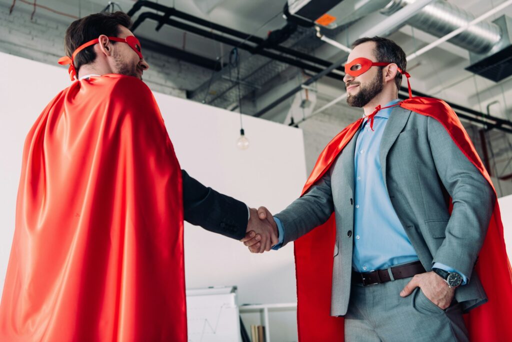 low angle view of handsome super businessmen in masks and capes shaking hands in office